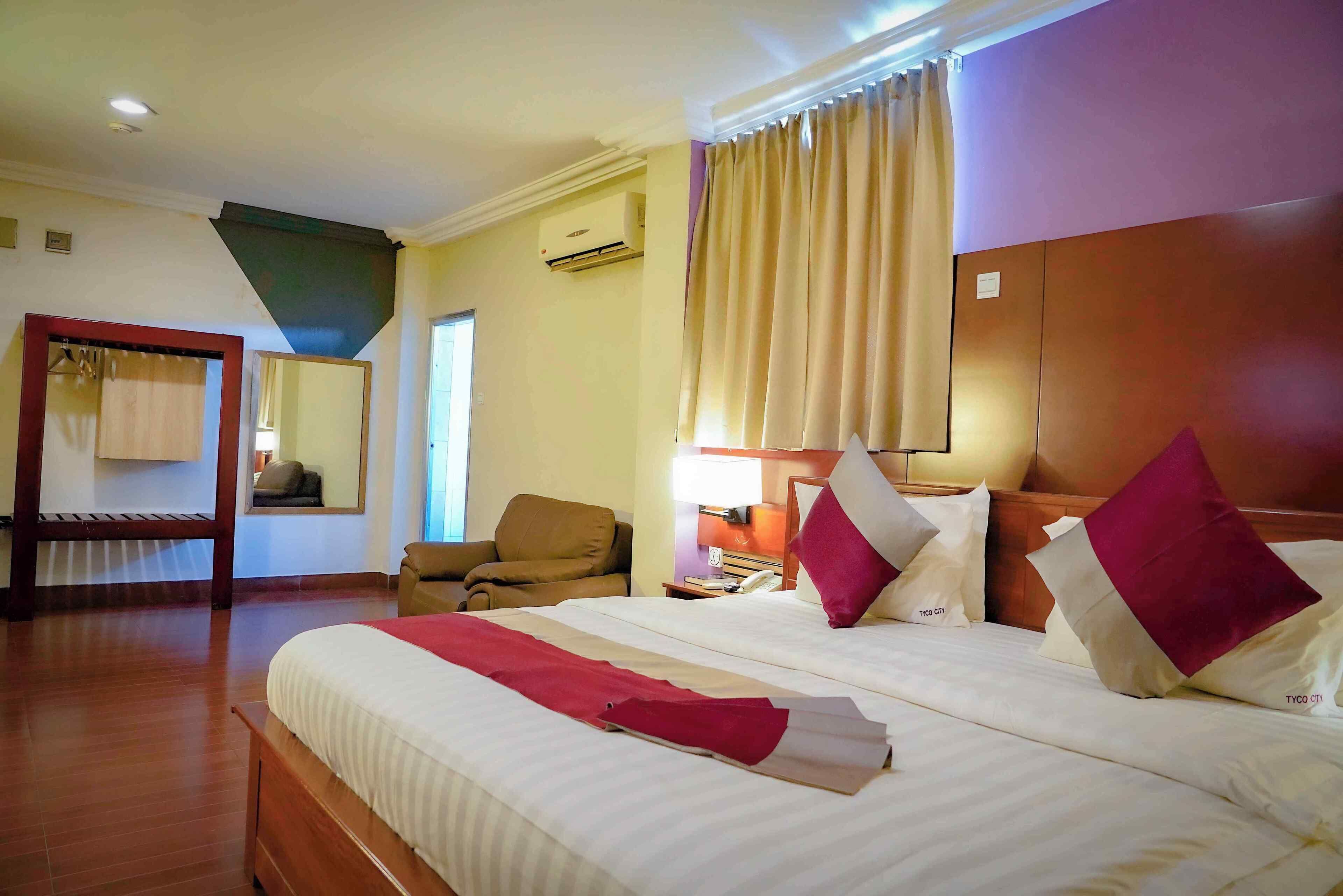 A hotel room with a large bed, a wooden bench, a chair, a bedside table with a lamp, and a window with curtains.