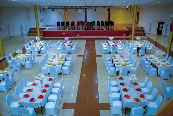 Un salón de banquetes con varias mesas redondas y sillas, un escenario con un telón rojo, y una escalera que conduce a un nivel superior.