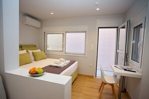 A compact hotel room with a bed, a desk, and a chair. There is a bowl of fruit on the bed and a window with a view. - Emily in Athens - Central balcony flat (Athina)