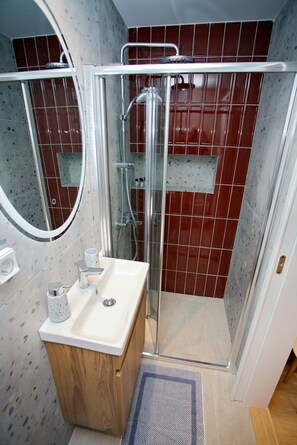 A bathroom with a glass shower enclosure, a sink with a mirror, and red-tiled walls. - Emily in Athens - Central balcony flat (Athina)