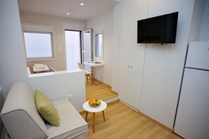 A compact living space with a sofa, a small table with fruit, a TV mounted on the wall, and a kitchenette area. - Emily in Athens - Central balcony flat (Athina)