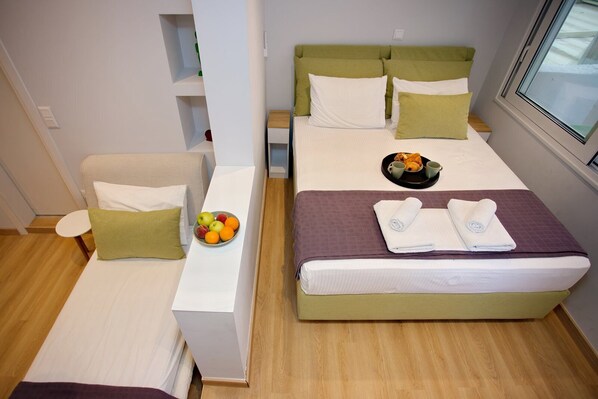 A compact hotel room with a bed, a small seating area, a bedside table with a fruit bowl, and a window with a view. - Emily in Athens - Central balcony flat (Athina)