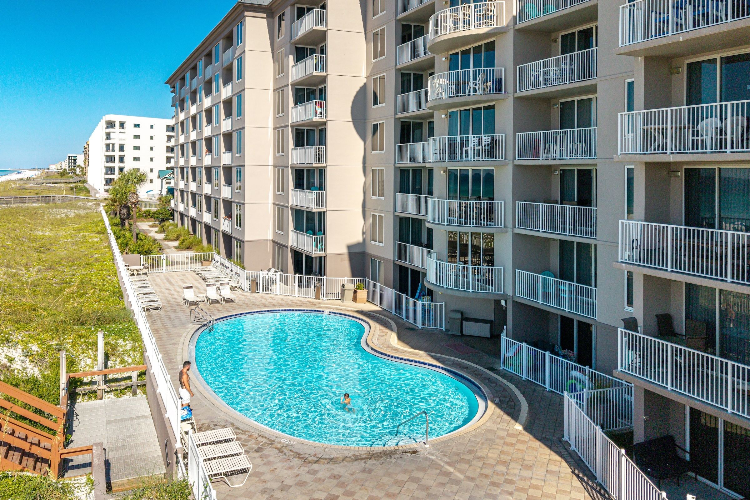 A multi-story hotel with a swimming pool, balconies, and a view of the beach.