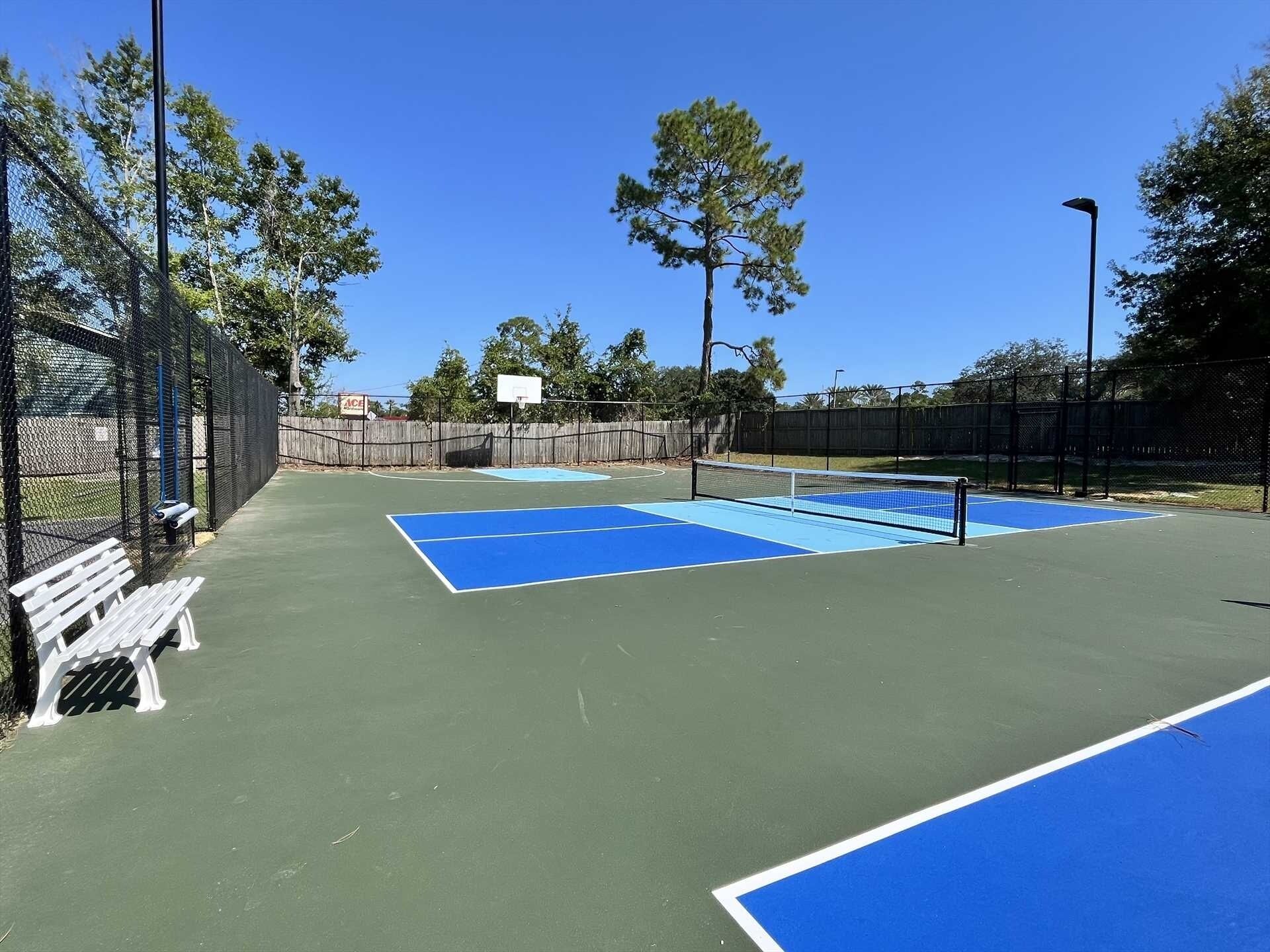 An outdoor tennis court with a basketball hoop, surrounded by a chain-link fence and trees.
