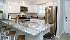 Una cucina moderna con mobili bianchi, un frigorifero in acciaio inossidabile, un forno a microonde integrato, un piano di lavoro in marmo e sgabelli da bar in legno.