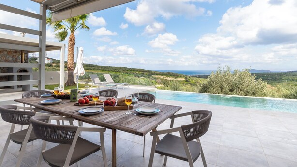 A wooden table set for a meal with a view of a swimming pool and landscape. - Villa Chryso (Platanias)