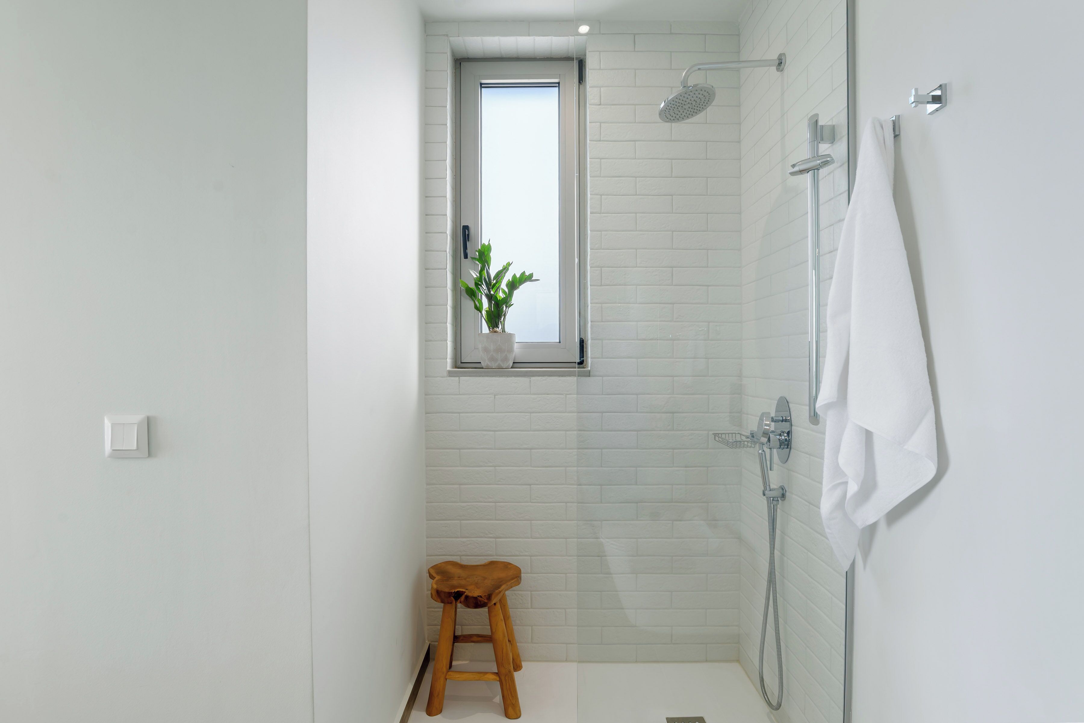 A small, modern bathroom with white tiles, a shower, a towel rack, and a window with a plant.