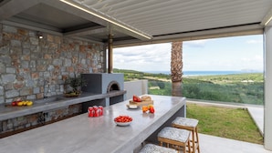 A modern kitchen with a stone wall, a built-in oven, and a large window overlooking a scenic landscape. - Villa Chryso (Platanias)