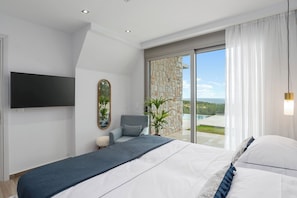 A modern bedroom with a large bed, a flat-screen TV, a chair, and a view of the outdoors. - Villa Chryso (Platanias)