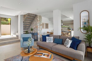 A modern living room with a stone staircase, a grey sofa with blue cushions, and a wooden coffee table with magazines. - Villa Chryso (Platanias)