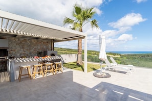 An outdoor kitchen with stone walls, a grill, and bar stools. There are lounge chairs and a view of the landscape. - Villa Chryso (Platanias)