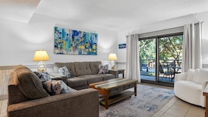 Un soggiorno con un divano grigio, un tavolino in legno e due lampade. Al muro, un quadro astratto di grandi dimensioni e vista sull'esterno attraverso una porta scorrevole in vetro.