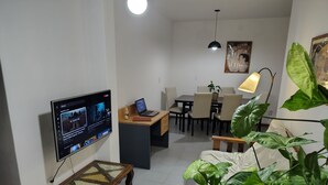 En moderne stue med et fladskærms-tv, en bærbar computer, et spisebord og stole. Der er et sofabord med glasplade og en lille plante.