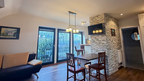 A modern living room with a dark sofa, wooden dining table, and chairs. There is a stone accent wall and a TV mounted above the fireplace. - Fully furnished flat in a lovely villa (Bergisch Gladbach)