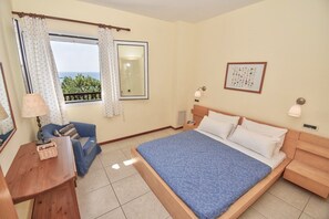 A hotel room with a bed, a chair, a desk, a lamp, a picture frame, and a window with curtains. - Hippo One Bedroom Apartment (Pelekas)