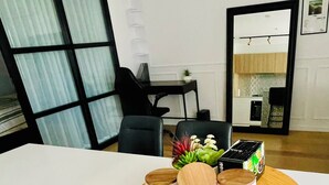 Moderni olohuone, jossa on suuri ikkuna, ruokailutila pöytineen ja tuoleineen sekä avoimen oven kautta näkyvä keittiö.