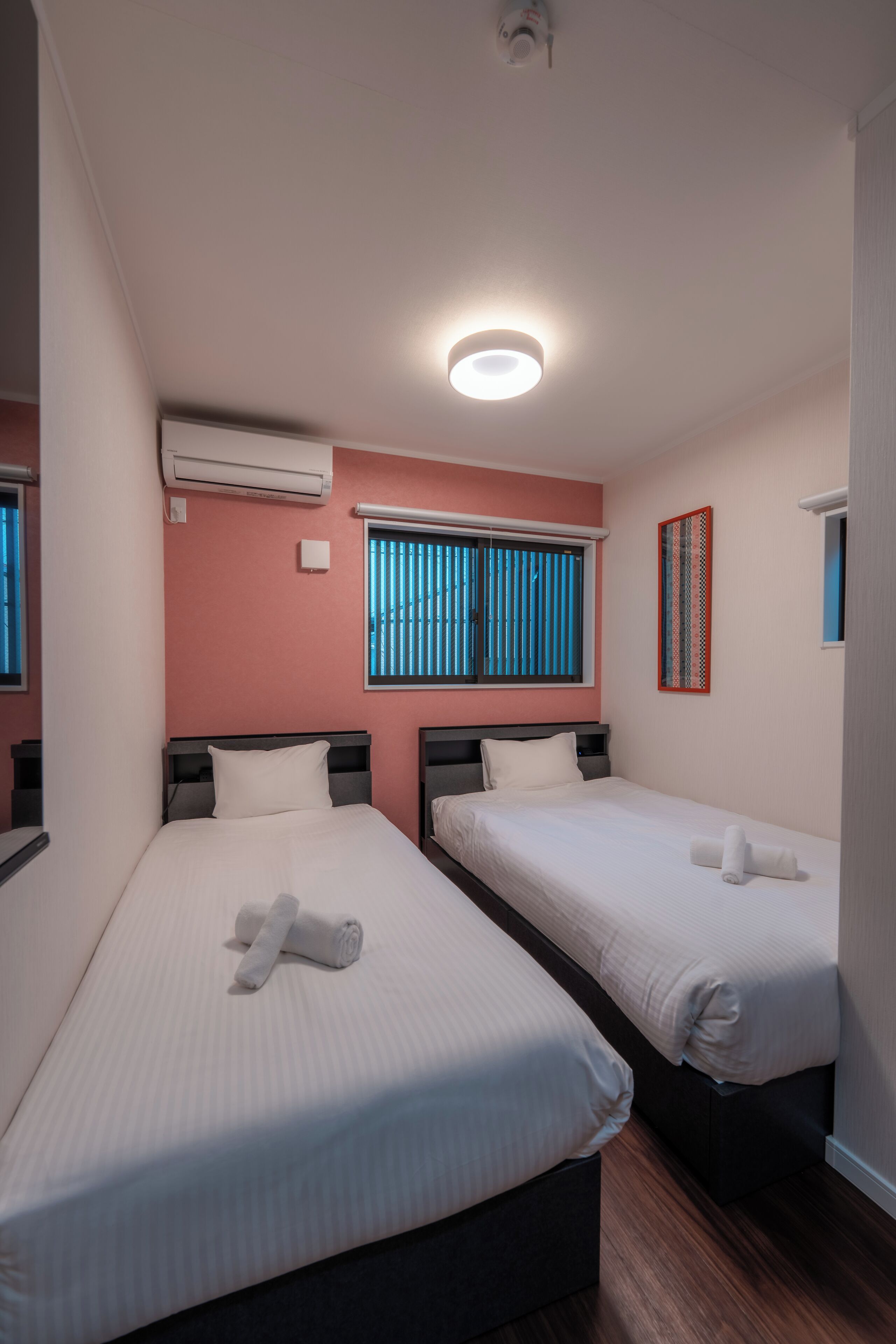 A hotel room with two single beds, a ceiling light, an air conditioning unit, and a window with blinds.