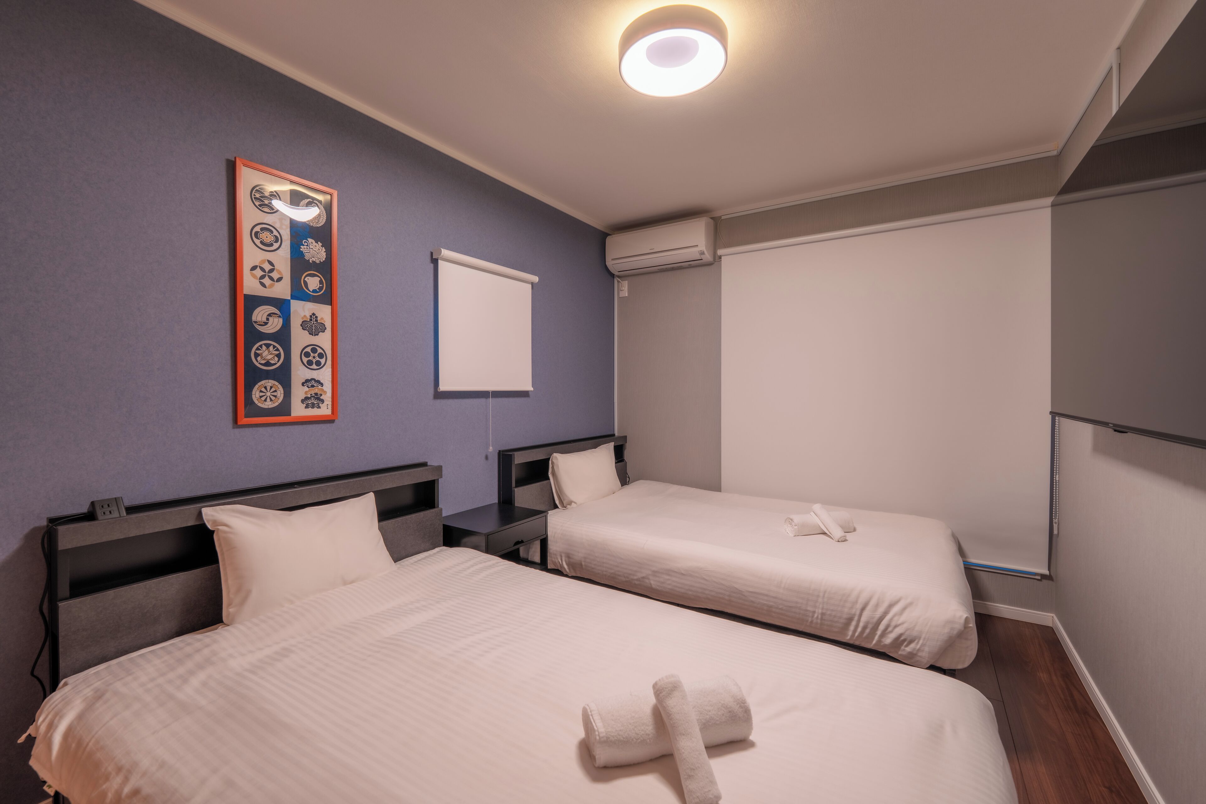 A hotel room with two beds, a wall-mounted air conditioner, a projector screen, and a framed artwork on the wall.
