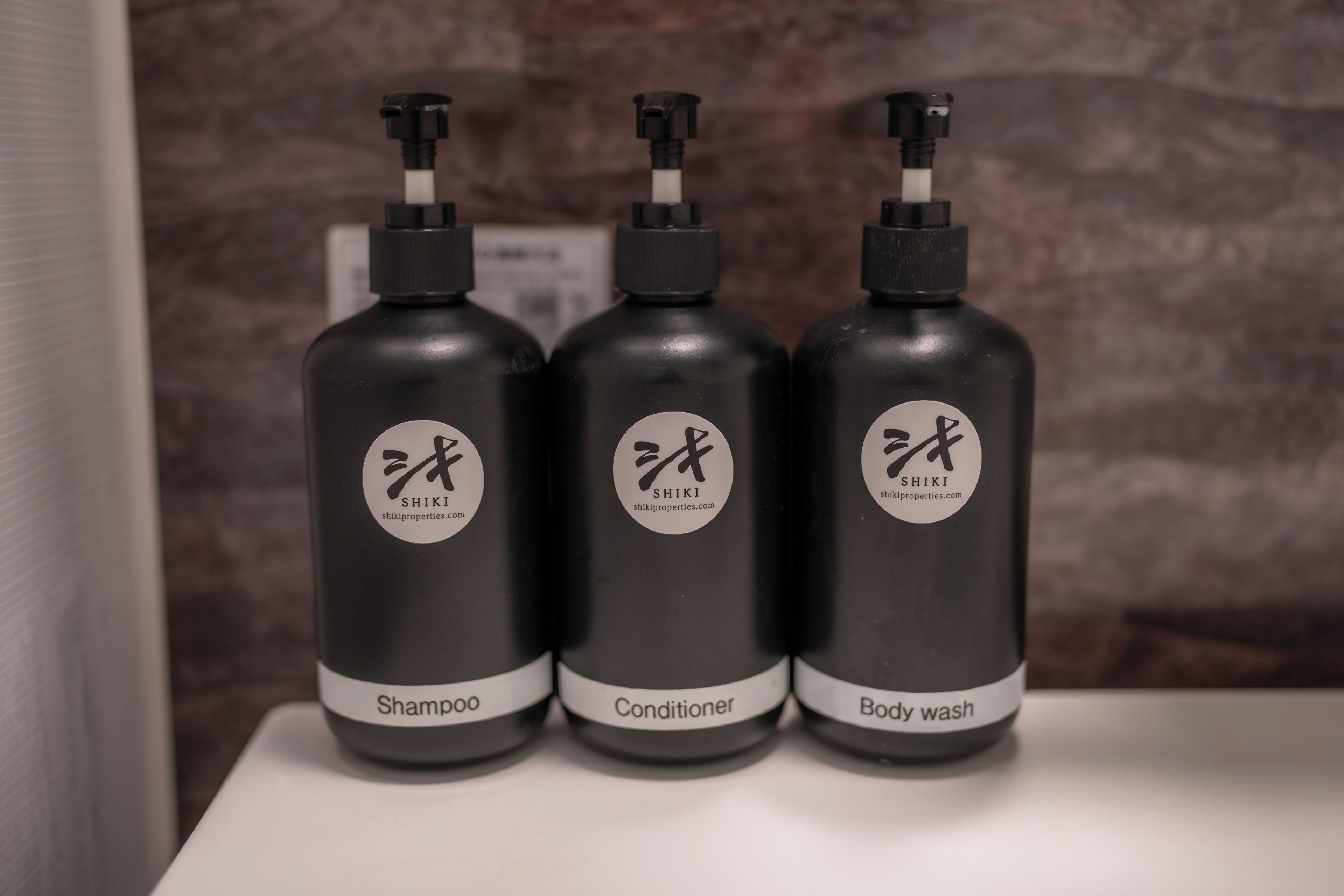 Drei schwarze Flaschen mit Pumpspendern, beschriftet mit 'Shampoo', 'Conditioner' und 'Body Wash', auf einer weißen Oberfläche.