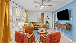 A living room with a brown sofa, orange chairs, a ceiling fan, and a TV mounted on the wall. - 5RM2BA,Game Rm,FirePit,Golf,BBall,Near Parris Island (Beaufort)