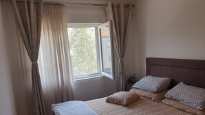 Un dormitorio con una cama, dos almohadas, una ventana con cortinas y una mesita de noche con un reloj.