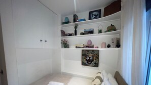 Quarto bem arrumado com uma cama, uma estante com livros e objetos decorativos, e uma janela com cortinas.
