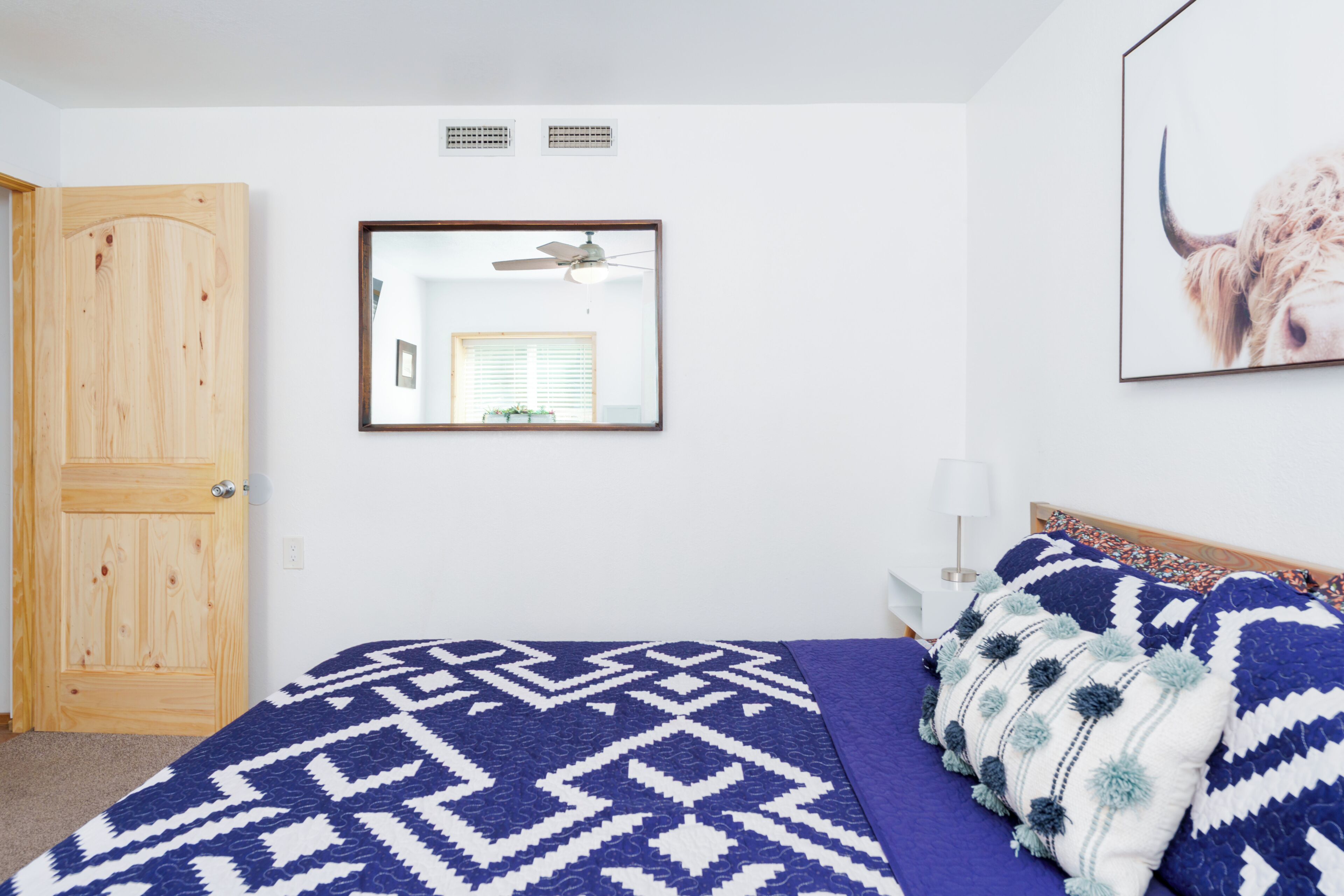Una camera da letto con una porta in legno, un letto con copriletto decorato, una ventola a soffitto, uno specchio e un quadro appeso al muro.
