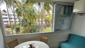 A room with a round dining table, four wooden chairs, a teal sofa, and a large window with a view of palm trees and a building. - Quiet, functional apartment - ideal for couples/families (Saint-Paul)