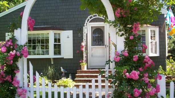 Una casa con valla blanca de piquetes, una puerta principal y plantas florecientes de color rosa.