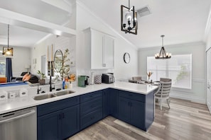 A modern kitchen with dark blue cabinets, white countertops, and a large island. There are various kitchen appliances and a dining area with a chair. - Lakefront Home w/ Private Pool & Scenic Views (Brandon)