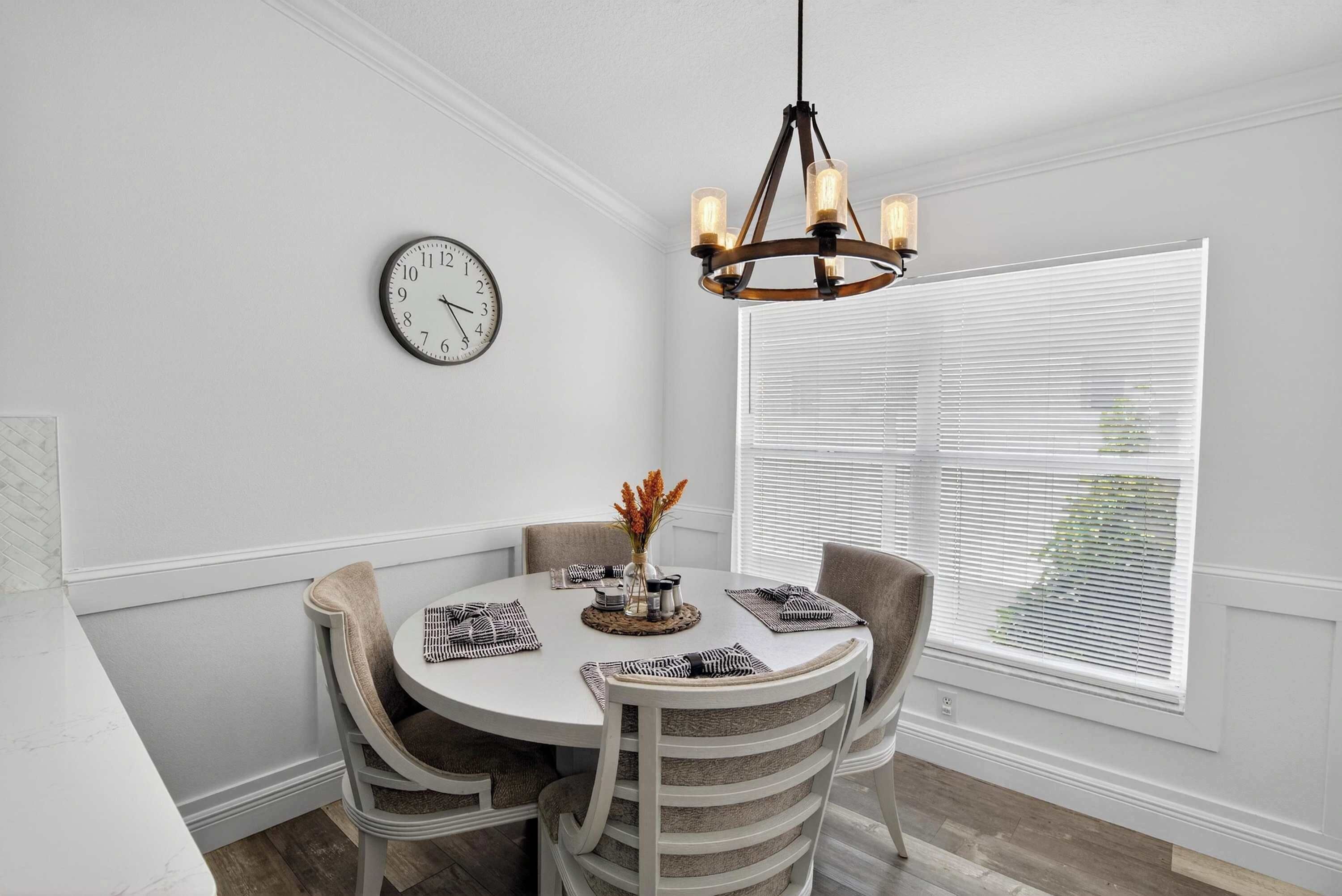 A dining area with a round table, four chairs, a chandelier, a wall clock, and a window with blinds.