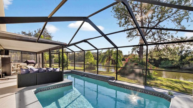 A swimming pool with a glass enclosure, a seating area with cushions, and a view of a lake and trees. - Lakefront Home w/ Private Pool & Scenic Views (Brandon)