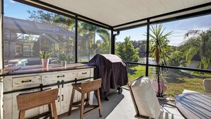 A covered outdoor area with a wooden table and chairs, a grill, and a view of a house and greenery. - Lakefront Home w/ Private Pool & Scenic Views (Brandon)