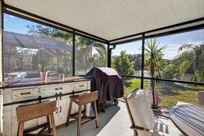 A covered outdoor area with a wooden table and chairs, a grill, and a view of a house and greenery. - Lakefront Home w/ Private Pool & Scenic Views (Brandon)