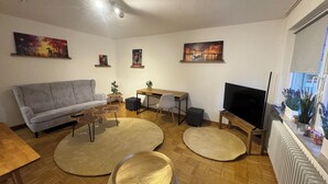 Un salón con un sofá gris, una mesa de centro de madera, alfombras redondas, un escritorio con una silla, un televisor y obras de arte en las paredes.