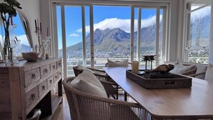 Espaço de refeições com móveis de vime, uma mesa de madeira e vista para montanhas através de grandes janelas.