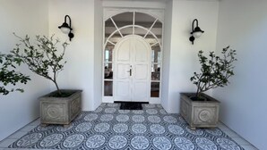 Uma entrada clara com piso de azulejos, duas plantas em vasos e uma porta branca com painel de vidro.