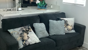 Ein modernes Wohnzimmer mit einem dunkelgrauen Sofa, einer Küchenzeile mit weißen Schränken, einer Edelstahl-Mikrowelle und einer Zimmerpflanze mit roten Blüten.