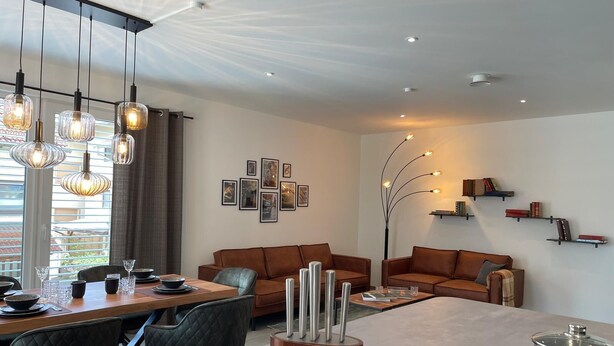 Een moderne woonkamer met een eethoek, met een zithoek, een salontafel en een eettafel met stoelen. Er zijn wandplanken met boeken en decoratieve objecten.