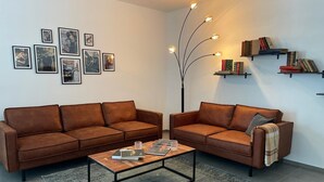 Een moderne woonkamer met twee bruine leren banken, een glazen salontafel en een vloerlamp. Er hangen ingelijste foto's aan de muur en er staan boekenrekken met boeken.