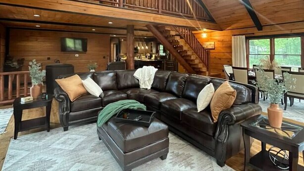 A spacious living room with a large sectional sofa, a coffee table, and a fireplace. There is a staircase leading to an upper level, and a large window providing natural light. - Meadow Ridge Lodge 4 Season Getaway (Altmar)