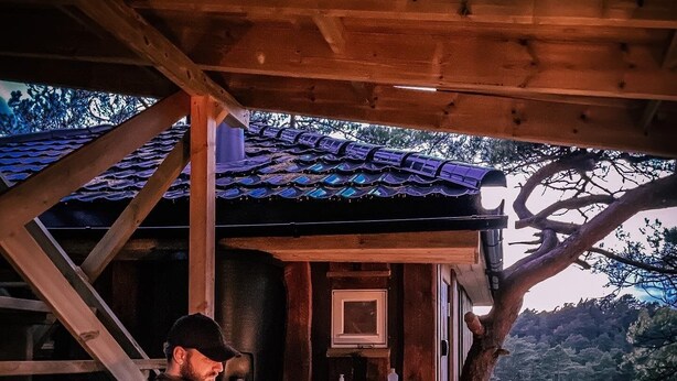 Un homme est assis à une table en bois, devant une bouteille de vin et une bougie, sous une structure en bois surmontée d’un toit en tuiles.