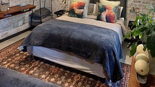 Een slaapkamer met een bakstenen muur, een bed met kleurrijke kussens, een hangstoel en een kleine tafel met een lamp.