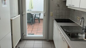 Een keuken met witte kastjes, een roestvrijstalen spoelbak en uitzicht door de deur op een terras met een tafel en stoelen.