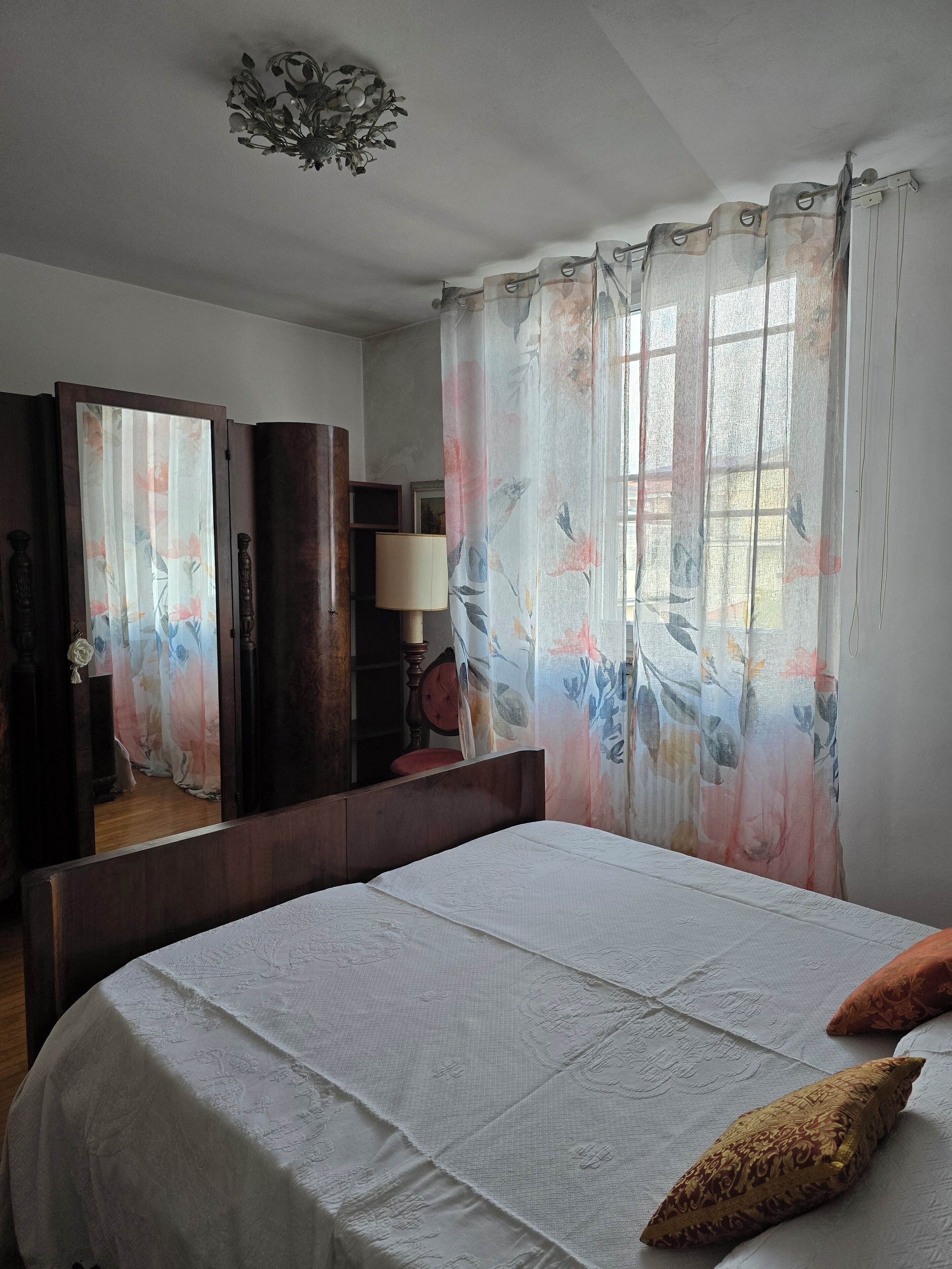 A bedroom with a large bed, a wooden wardrobe, a mirror, and a window with floral curtains.