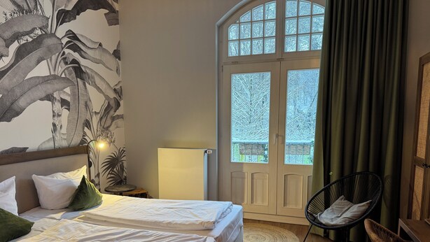 A bedroom with two beds, a chair, a lamp, and a view of a snowy landscape through a door. - Waldhotel Bad Essen  (Bad Essen)