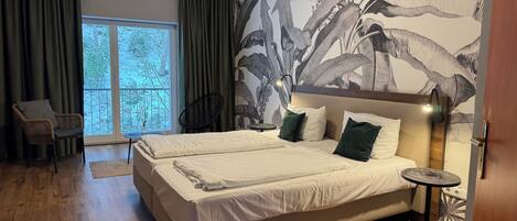 A modern bedroom with a large bed, a chair, a small table, and a view of greenery through the window.