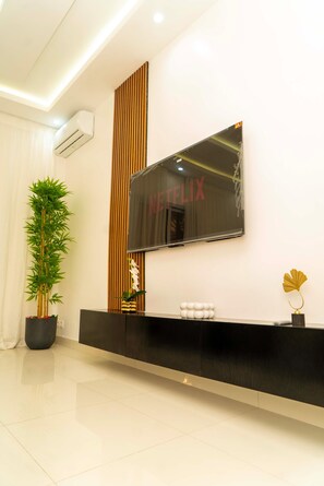 Una sala de estar moderna con un televisor de pantalla plana grande montado en la pared, un mueble de entretenimiento negro en la parte inferior, una planta alta en maceta y un jarrón decorativo.