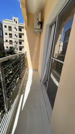 Un balcón estrecho con barandilla metálica, puerta corrediza de vidrio y un equipo de aire acondicionado instalado en la pared.