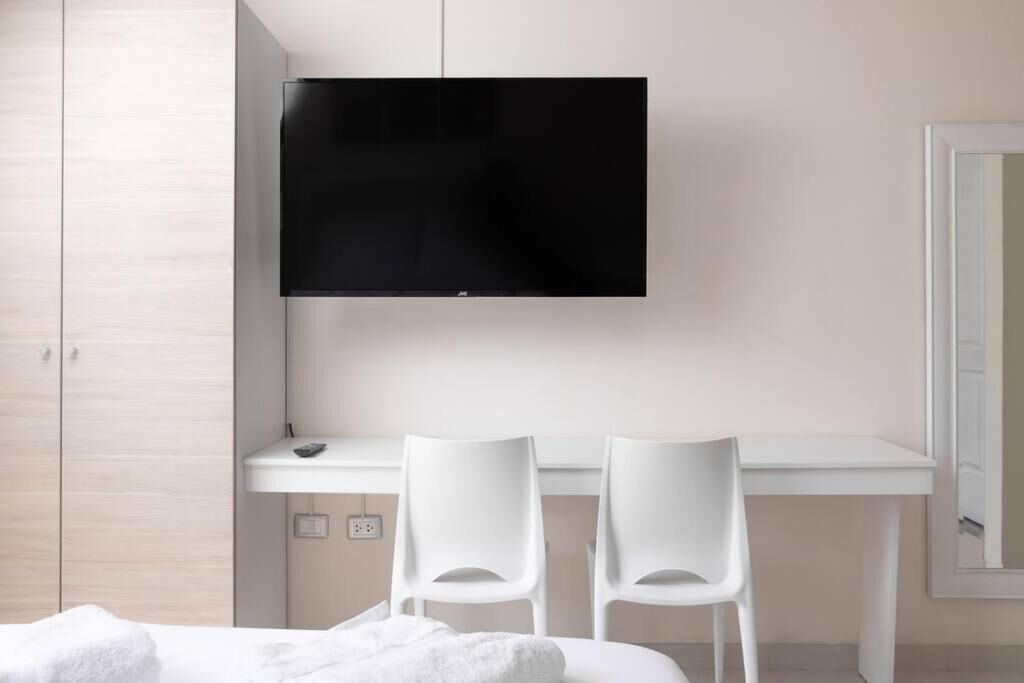 Una habitación de hotel moderna con una televisión de pantalla plana sobre un escritorio blanco, dos sillas blancas y una cama con una manta blanca.